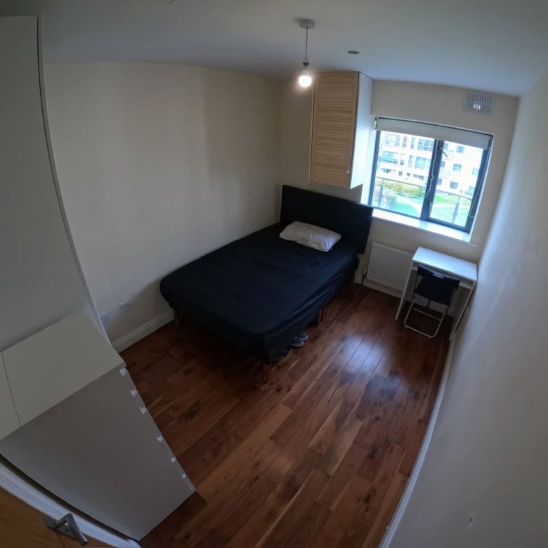 Small bedroom with bed, desk, window, and hardwood floor. Neutral colors.