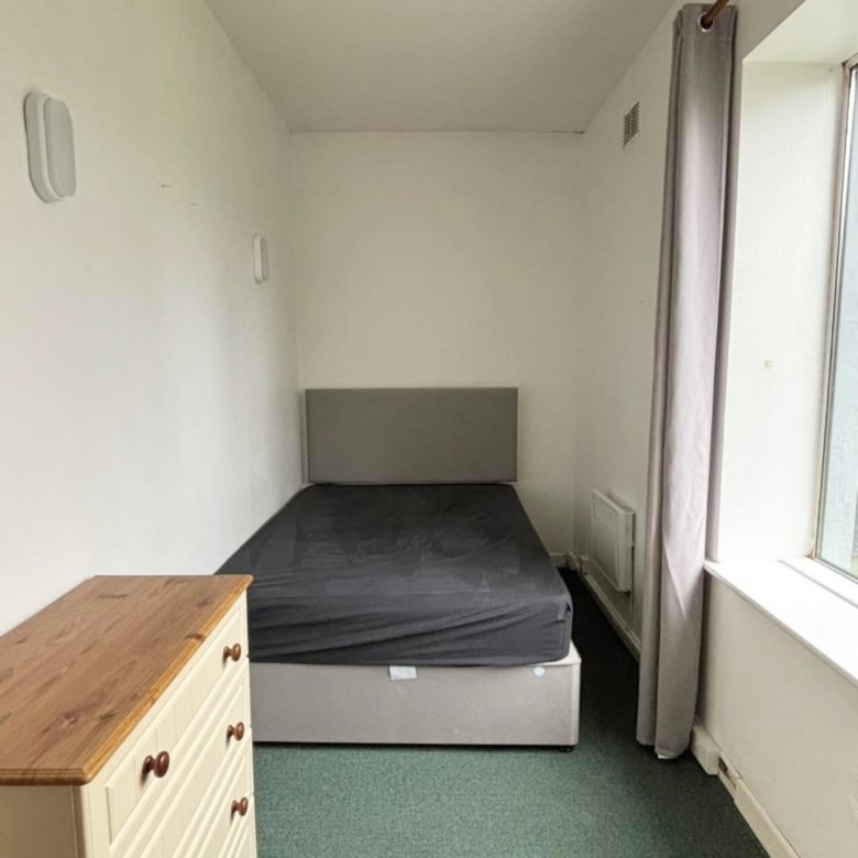 Small bedroom with bed, dresser, green carpet, and window with grey curtain.