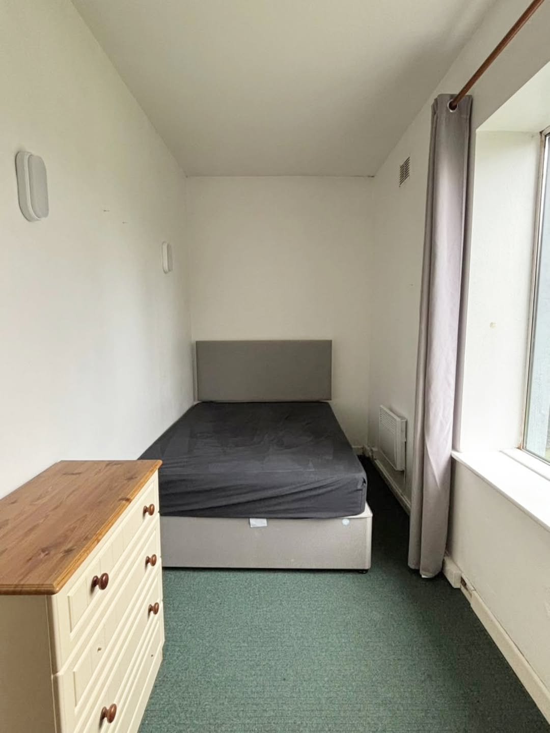 Small bedroom with bed, dresser, green carpet, and window with grey curtain.