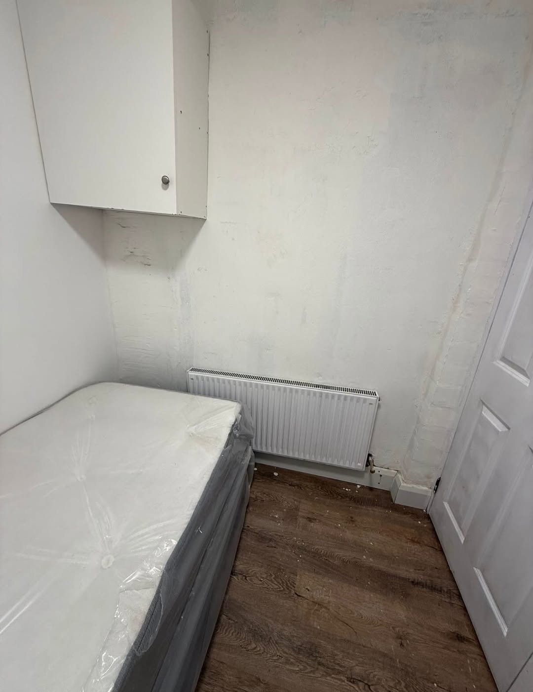 Small bedroom with bed, radiator, cabinet, and white walls. Minimalist interior design.