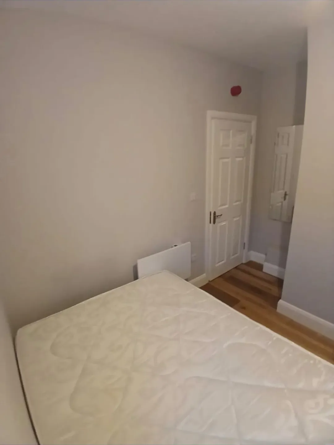 Small bedroom with bed, white walls, and wooden floor. Door and small cabinet visible.