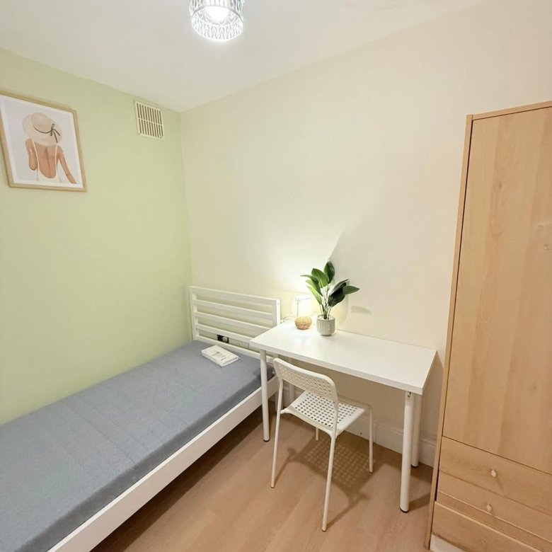 Small bedroom with single bed, desk, chair, and wardrobe. Light walls and wood floor. Plant on desk.