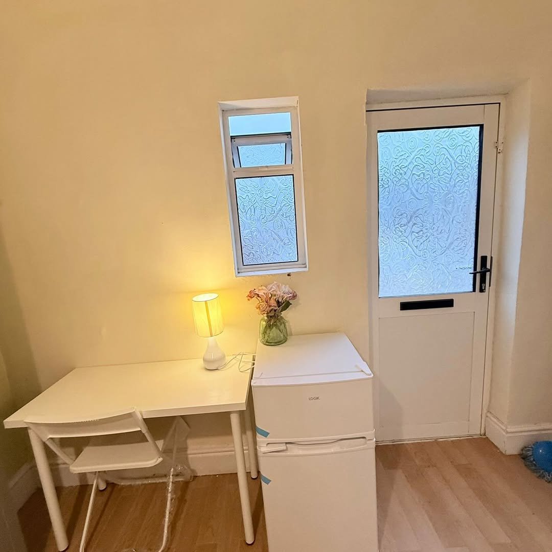 Small white desk and mini fridge in a room with a narrow window and door.