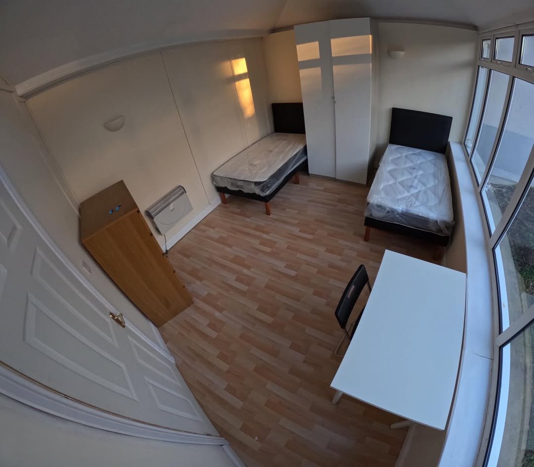 Small room with two beds, wardrobe, desk, and chair by window. Simple interior.