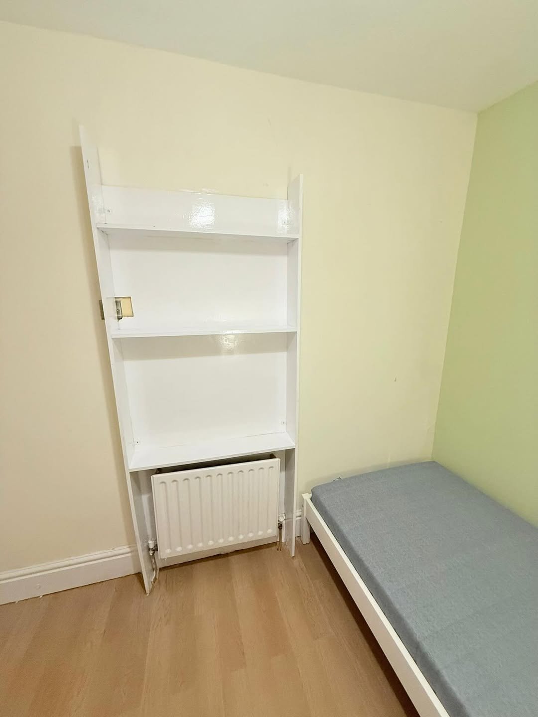 Small room with white shelving unit, radiator, and single bed against light yellow and green walls.