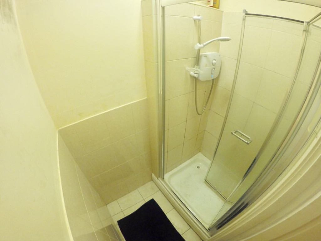 Small shower stall with glass door, white tile, and black bath mat in a bathroom setting.