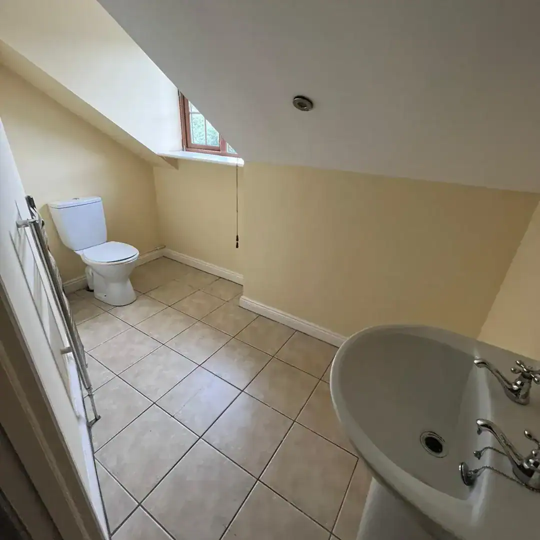 Attic bathroom with toilet, sink, tile floor, and small window under sloped ceiling. Neutral color scheme.