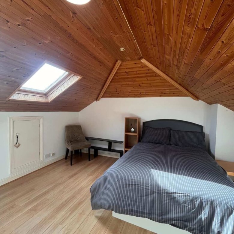 Attic bedroom with wood ceiling, skylight, gray bed, chair, and shelf. Light wood floor. Cozy space.