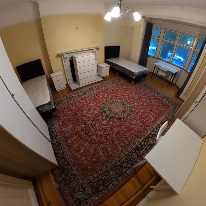 Bedroom with two single beds, red rug, fireplace, and desk near window. Bright lighting.