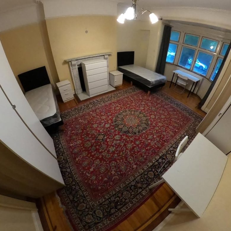 Bedroom with two single beds, red rug, fireplace, and desk near window. Bright lighting.