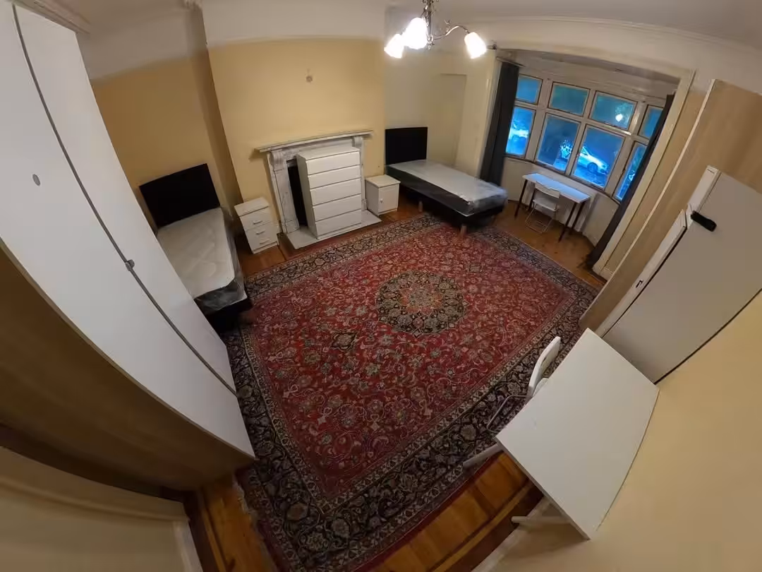 Bedroom with two single beds, red rug, fireplace, and desk near window. Bright lighting.