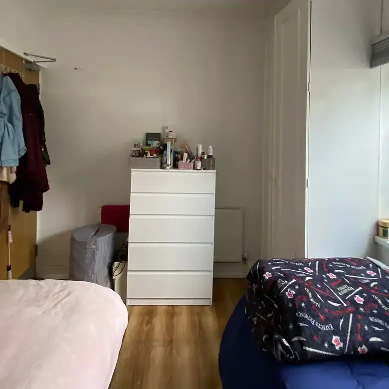 Bedroom with white dresser, wardrobe, bed, and clothes rack against a white wall. Natural light from window.