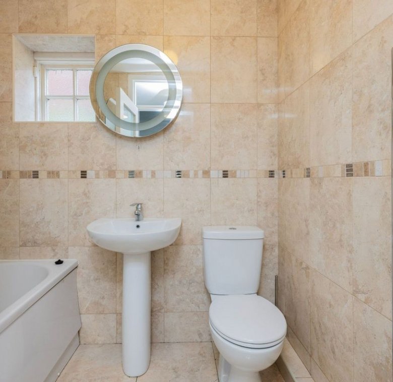 Beige tiled bathroom with toilet, sink, towel warmer, round mirror, and small window.