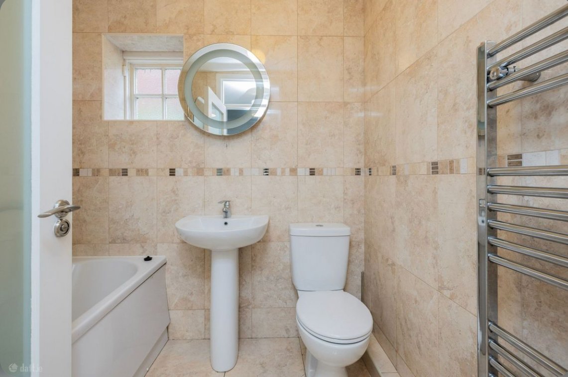 Beige tiled bathroom with toilet, sink, towel warmer, round mirror, and small window.