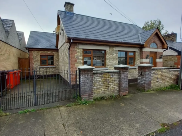 Brick bungalow house with slate roof, front garden wall, and black iron gate on a cloudy day.