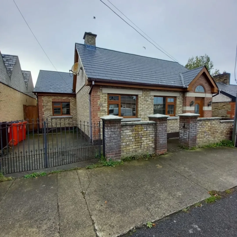 Brick bungalow house with slate roof, front garden wall, and black iron gate on a cloudy day.