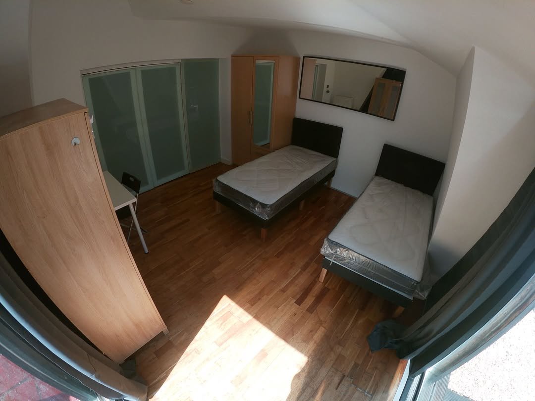 Bright bedroom with two single beds, wardrobe, desk, and large mirror. Hardwood floors and sloped ceiling.