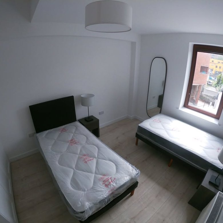 Bright bedroom with two single beds, white walls, wardrobe, mirror, and window showing an outside view.