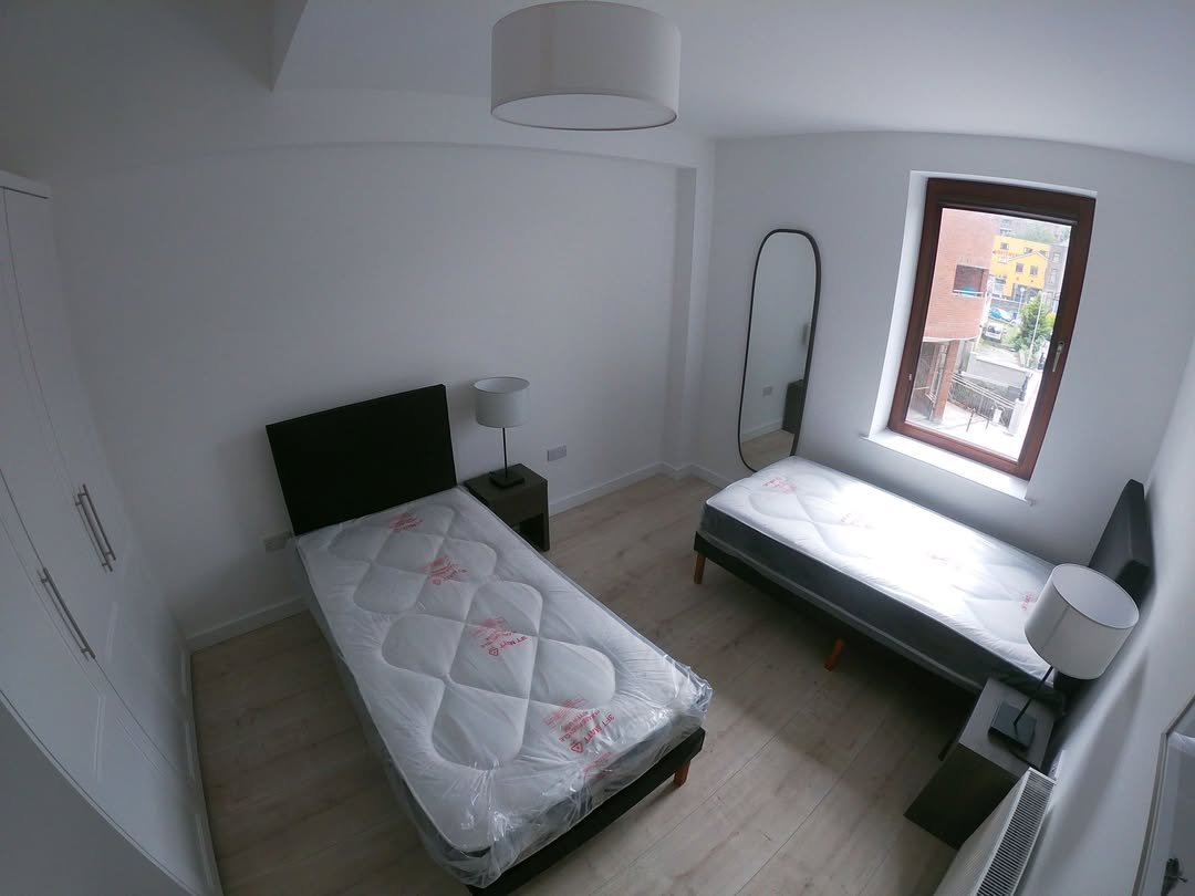Bright bedroom with two single beds, white walls, wardrobe, mirror, and window showing an outside view.