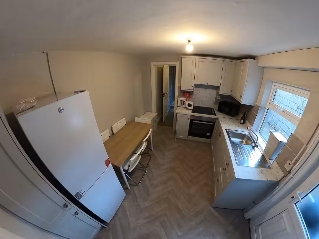 Bright kitchen with white cabinets, herringbone floor, dining table, and large fridge. Window provides natural light.