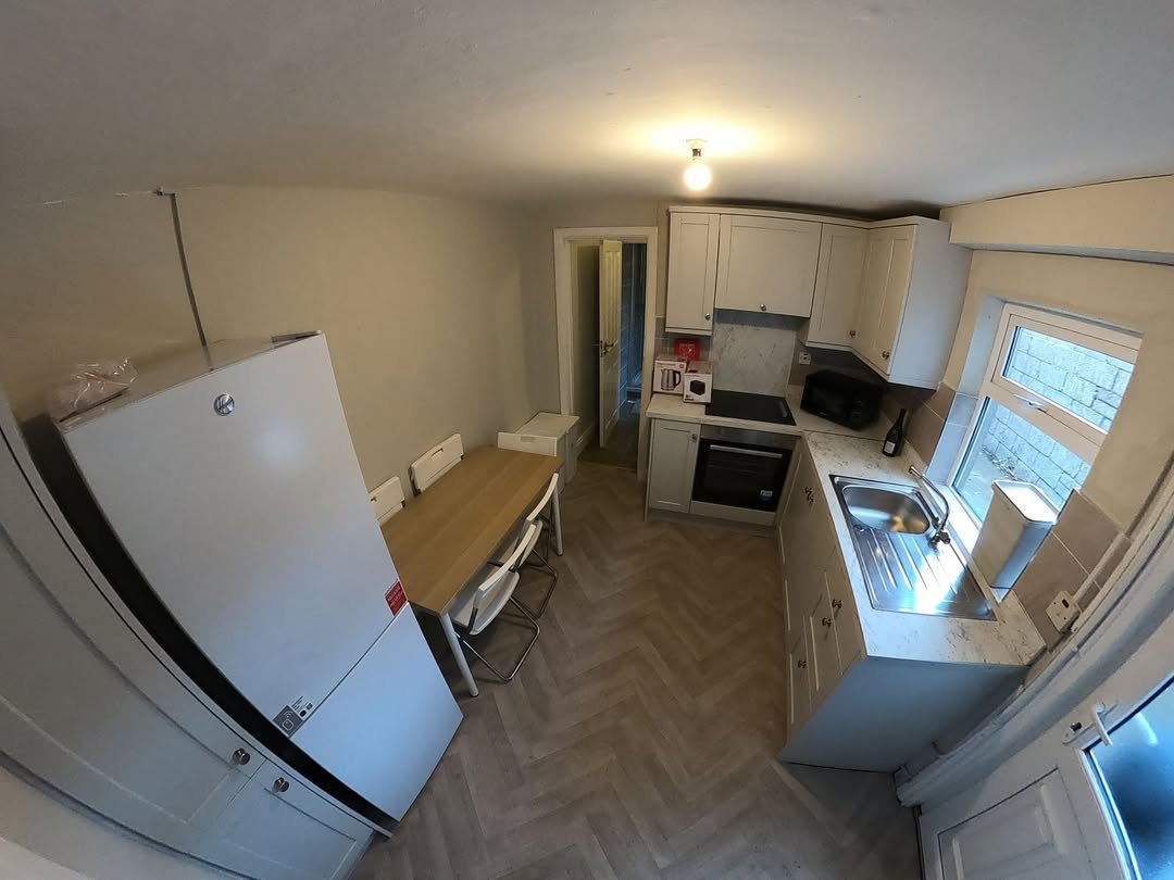 Bright kitchen with white cabinets, herringbone floor, dining table, and large fridge. Window provides natural light.