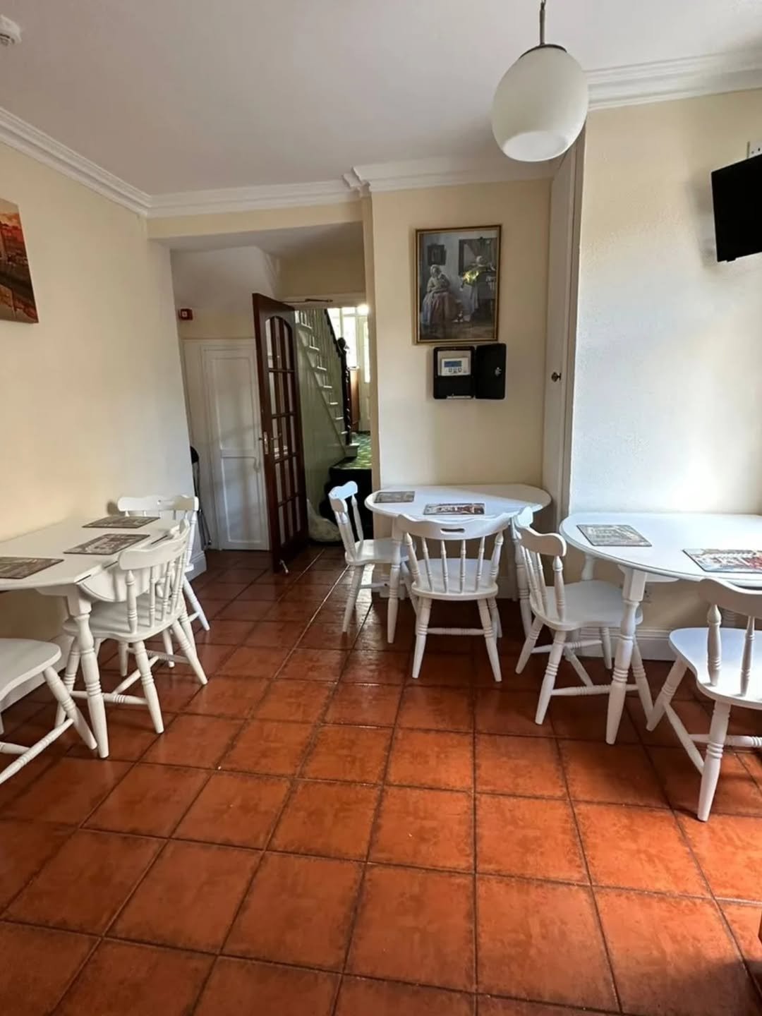 Cozy dining room with white tables, chairs, and terracotta tile flooring leading to a hallway and stairs.