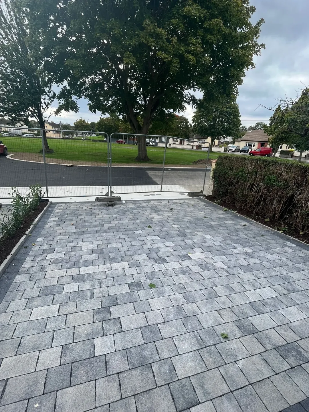 Gray brick driveway with temporary fencing and green trees in the background.