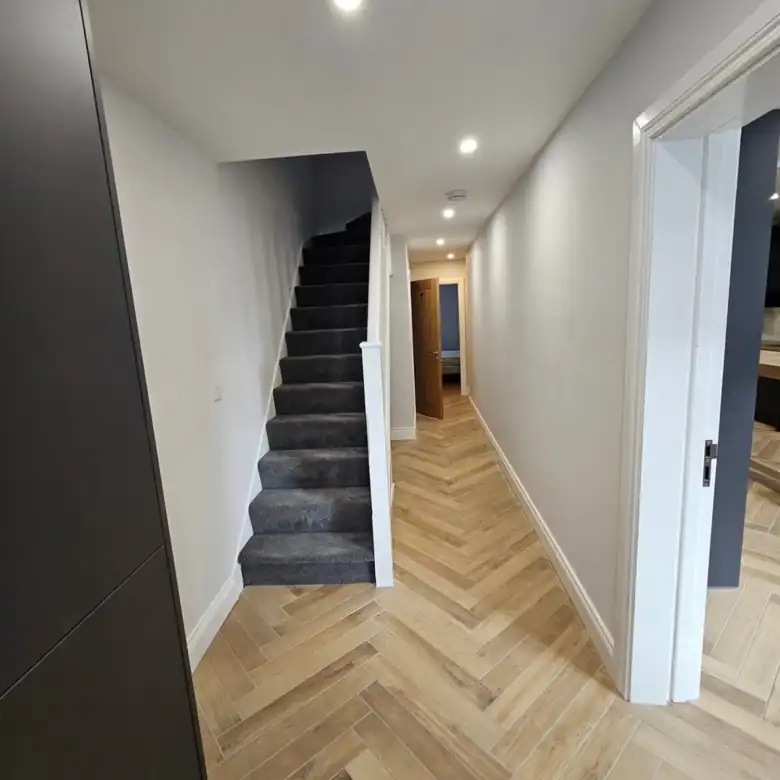 Hallway with herringbone wood floor, carpeted stairs, and white walls.