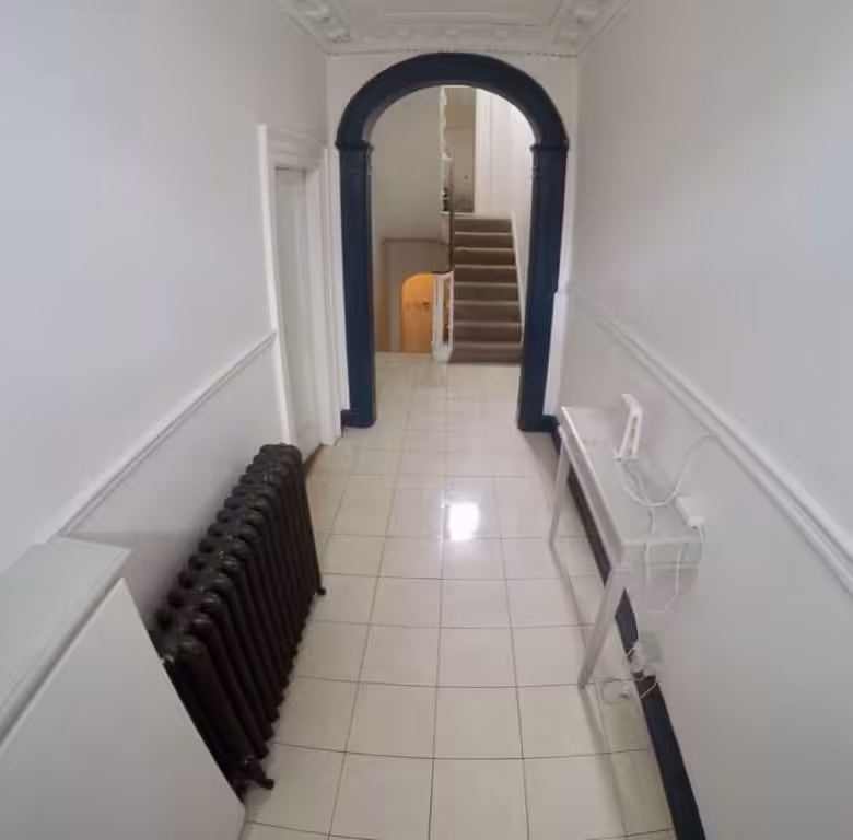 Hallway with radiator, table, and archway leading to stairs. White walls and tiled floor.