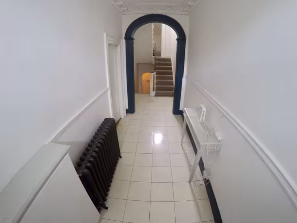 Hallway with radiator, table, and archway leading to stairs. White walls and tiled floor.