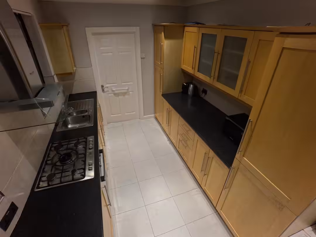 Kitchen with light wood cabinets, black countertops, stainless steel stove and sink, and white tile flooring.