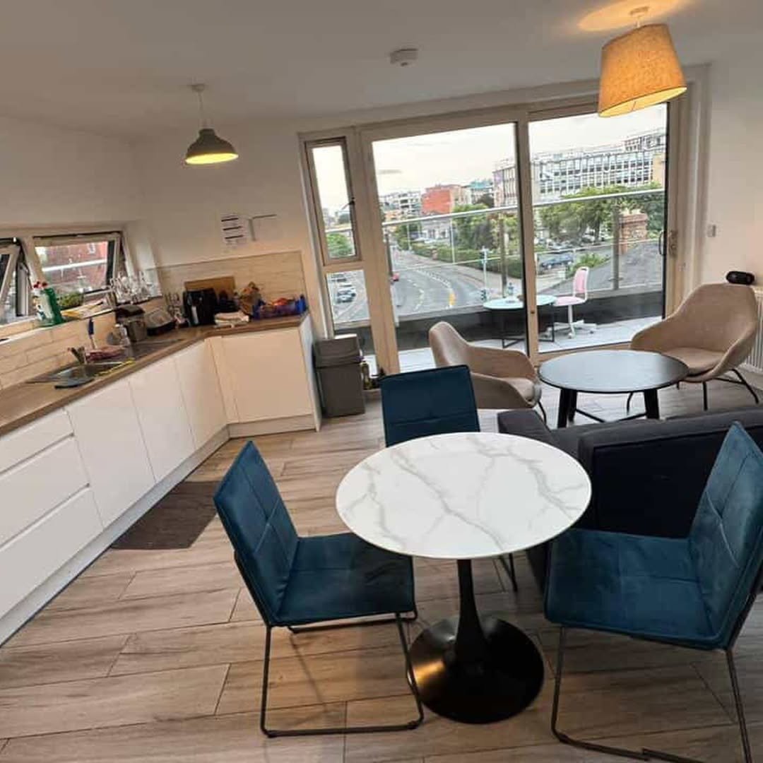 Modern apartment interior with dining area, kitchen, and balcony overlooking city. Neutral tones and blue accents.