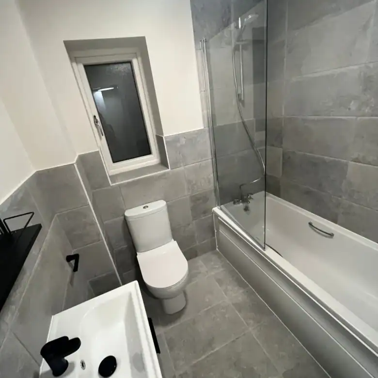 Modern bathroom featuring a toilet, sink, bathtub with glass screen, and gray tile walls and floor.