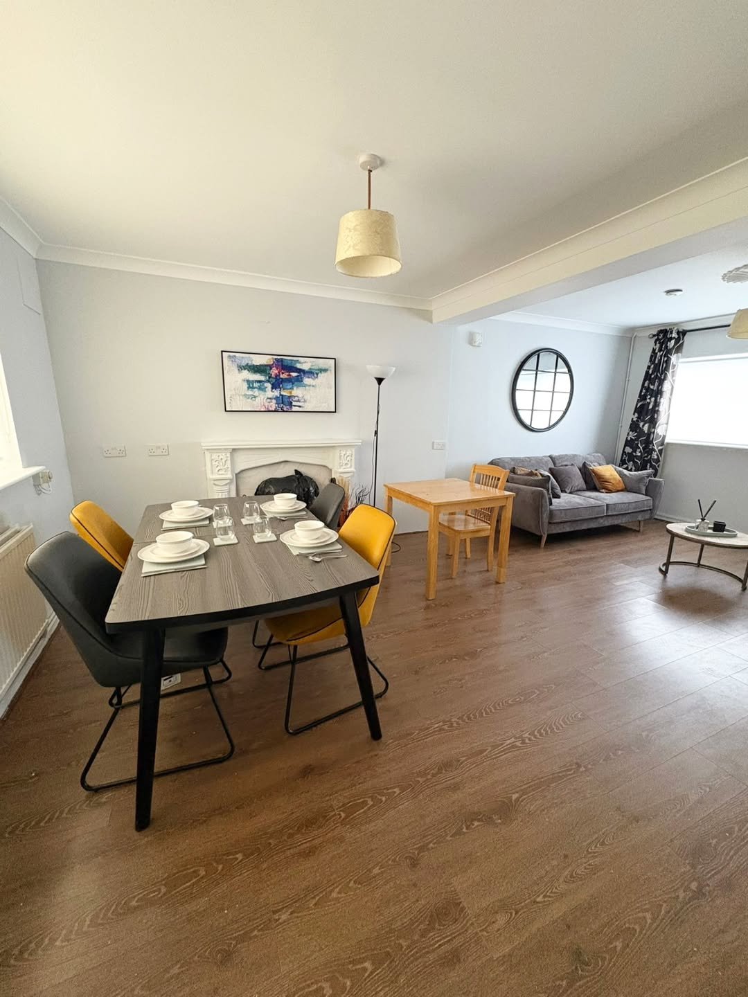 Modern living room with dining area, gray sofa, wood floors, and table with yellow chairs.