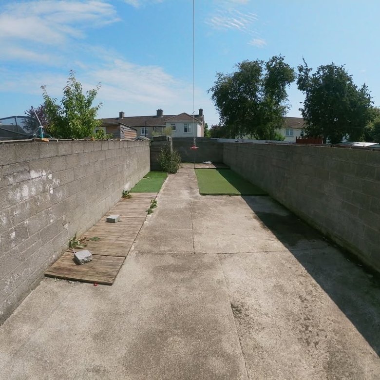 Narrow backyard with concrete patio, cinder block walls, and patches of artificial grass. Blue sky above.