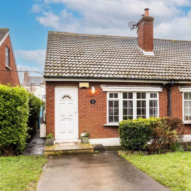 Red brick bungalow with white door and windows, green bushes, and a chimney.