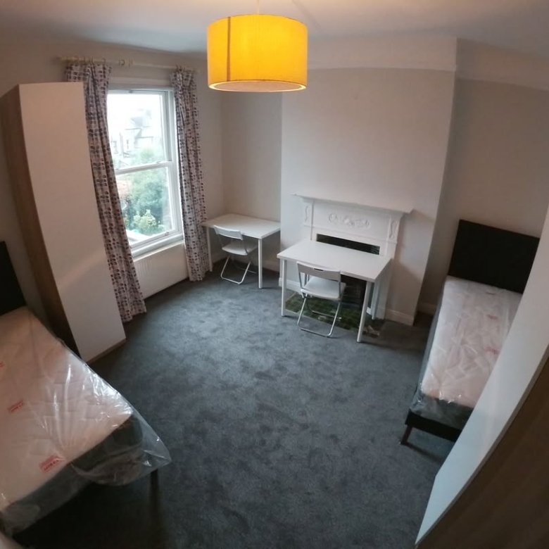 Shared bedroom with two beds, desks, wardrobes, grey carpet, and natural light from a window.