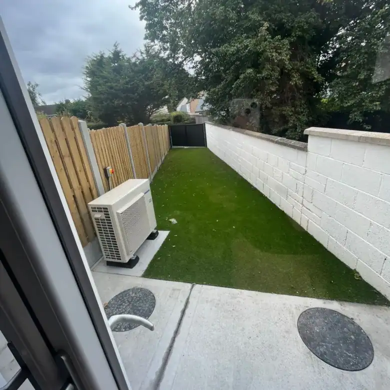 Small backyard with artificial grass, white wall, wooden fence, and air conditioning unit.