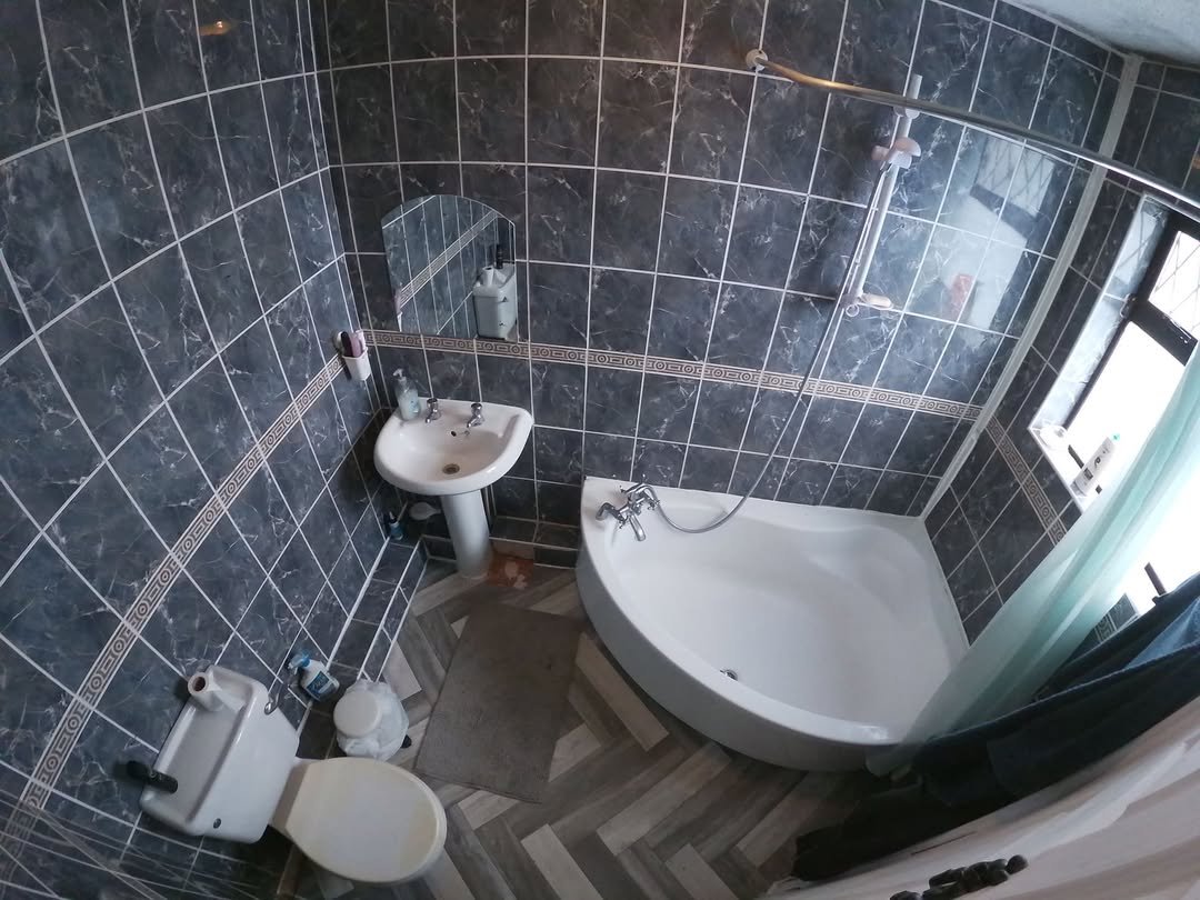 Small bathroom with gray tiles, pedestal sink, toilet, and corner bathtub with shower.