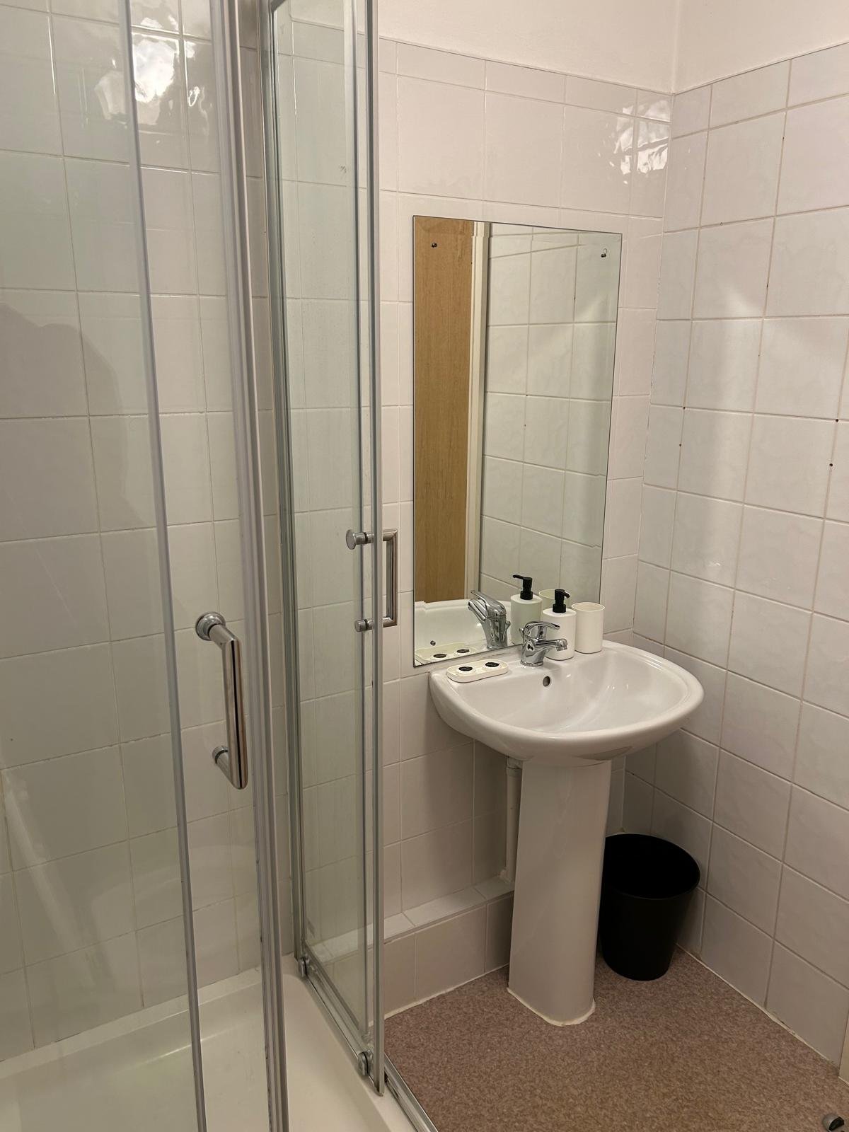 Small bathroom with shower, pedestal sink, and mirror in a tiled room.