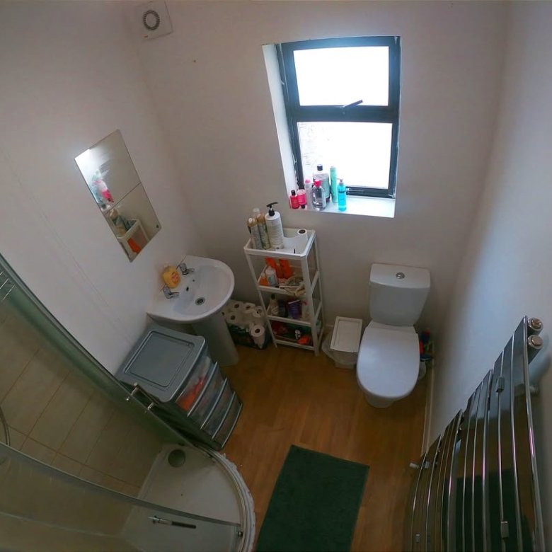 Small bathroom with shower, toilet, sink, and window. White walls and light wood floor.
