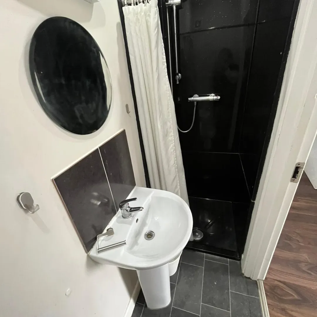 Small bathroom with white sink, black shower stall, round mirror, and gray tile flooring.