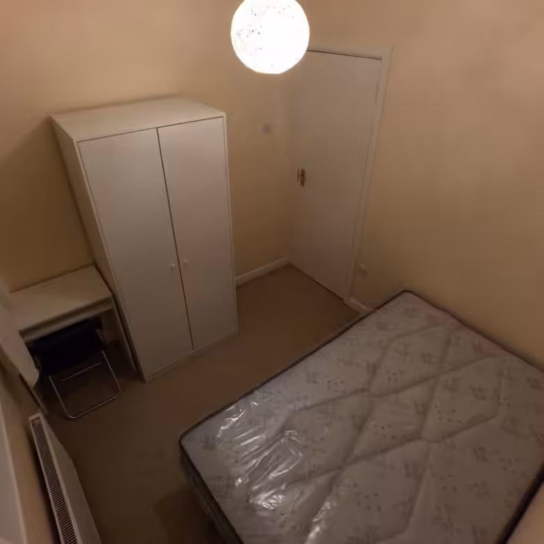 Small bedroom interior: bed, wardrobe, desk, and radiator under a bright ceiling light.