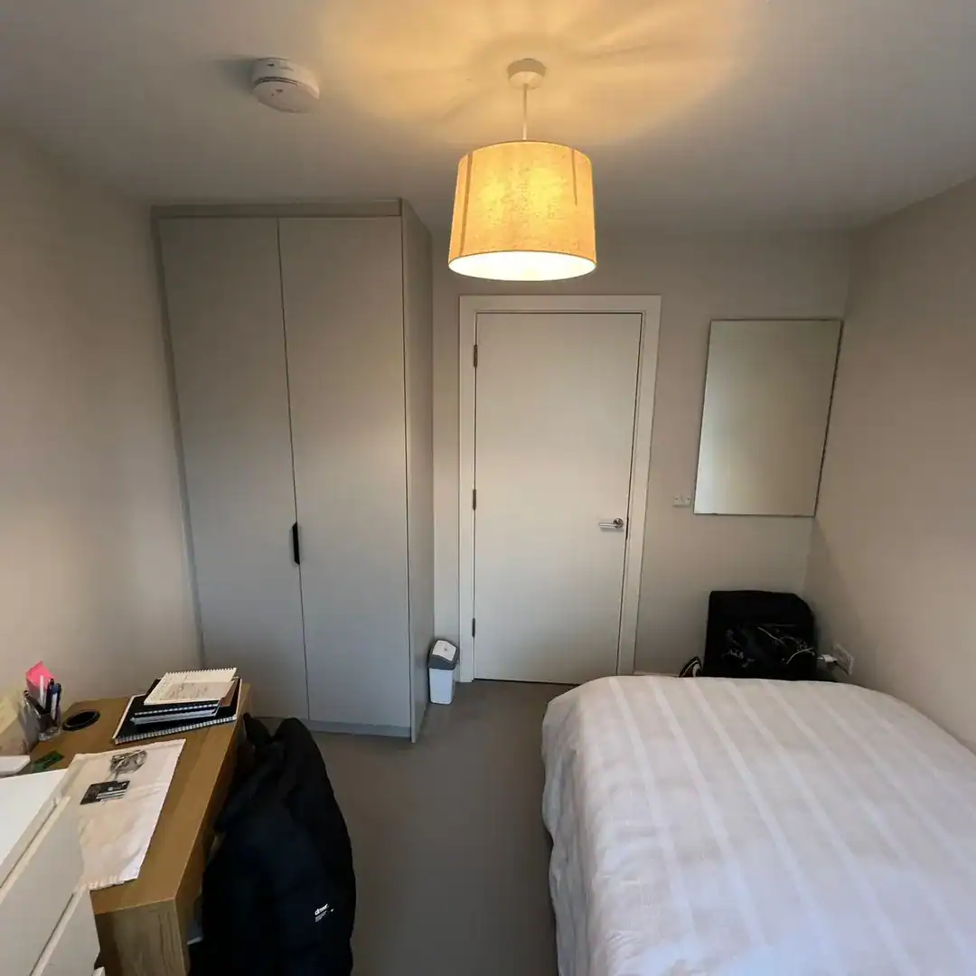 Small bedroom interior with bed, wardrobe, desk, door, and mirror.