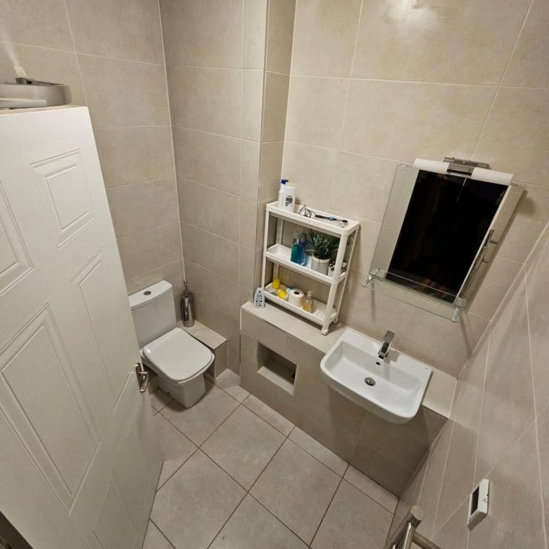 Small beige tiled bathroom with toilet, sink, and white storage shelf.