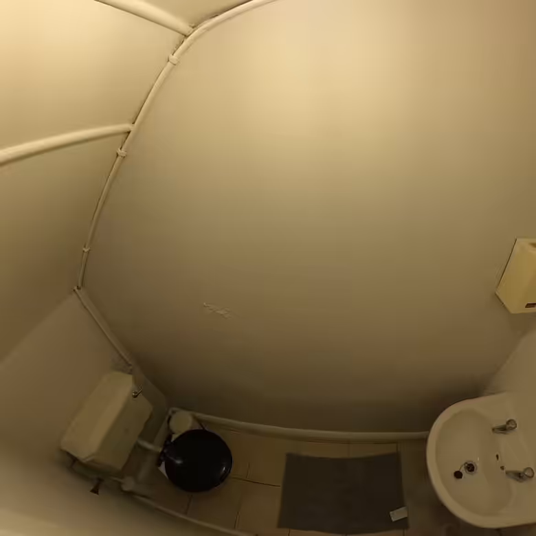 Small, curved bathroom with toilet, sink, and pipes. White walls and tile floor.