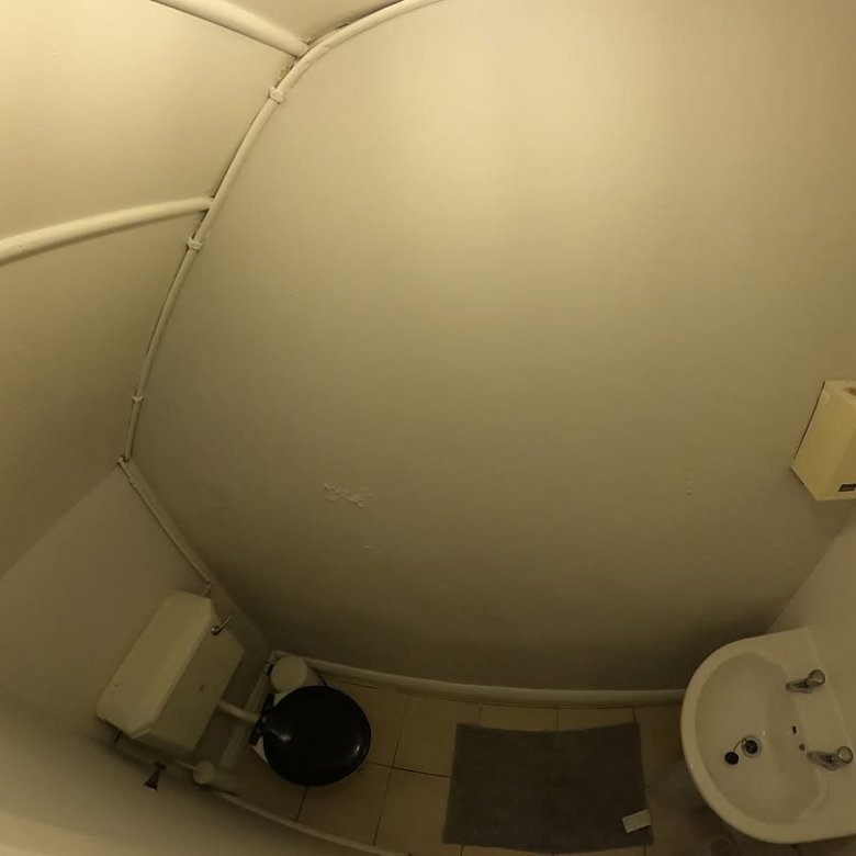 Small, curved bathroom with toilet, sink, and pipes. White walls and tile floor.