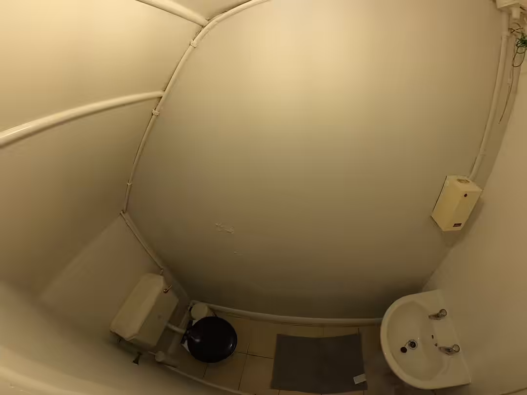 Small, curved bathroom with toilet, sink, and pipes. White walls and tile floor.