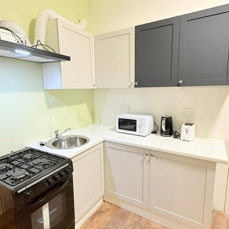 Compact kitchen with black gas stove, sink, microwave, kettle, toaster, and cream/gray cabinets under a vent hood.