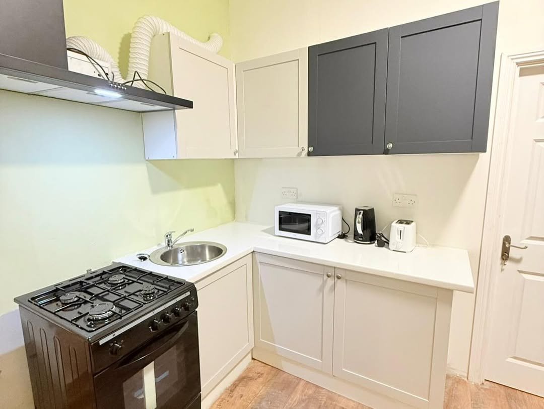 Compact kitchen with black gas stove, sink, microwave, kettle, toaster, and cream/gray cabinets under a vent hood.